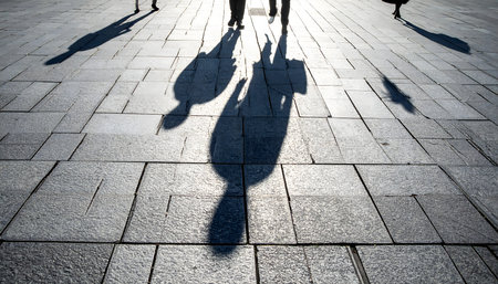 Low-angle perspective of people's feet and long shadows walking across a brightly lit gray tiled public squareの素材