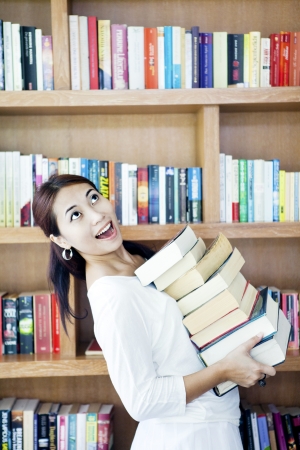 Young attractive asian woman holding a pile of books in front of bookshelfの写真素材
