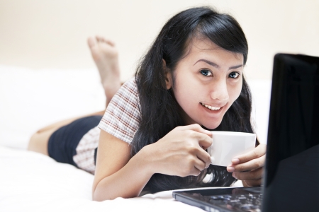 Smiling sweet asian woman with laptop and cup of coffee on the bedの写真素材
