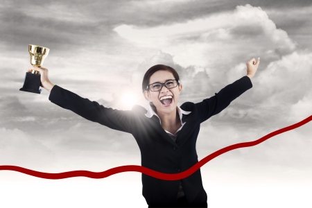 Businesswoman celebrates her winning and running thru the ribbon while holding trophyの写真素材