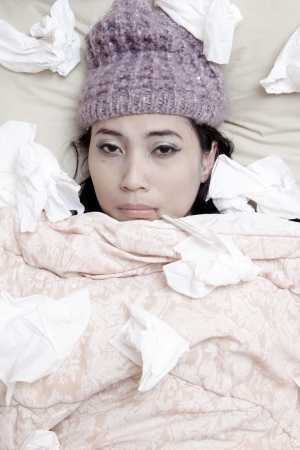 Woman is laying on her bed, wearing beany due to flu winterの写真素材