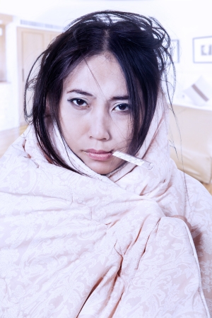 Woman having fever wrapped in blanket while biting a thermometer in her mouthの写真素材