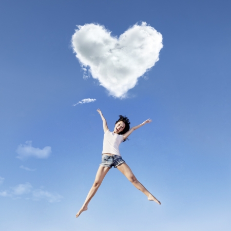 Beautiful girl is jumping under a heart shape cloudの写真素材