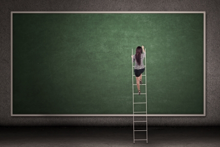 Businesswoman is climbing a ladder in front of a blackboardの写真素材