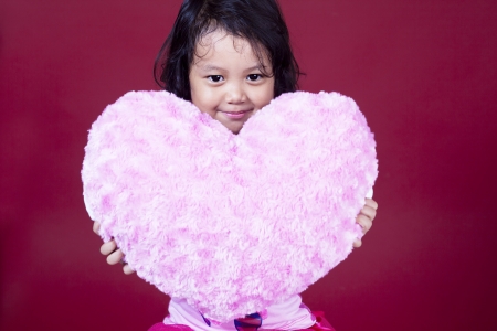 Cute girl is holding heart pillow on pink backgroundの写真素材
