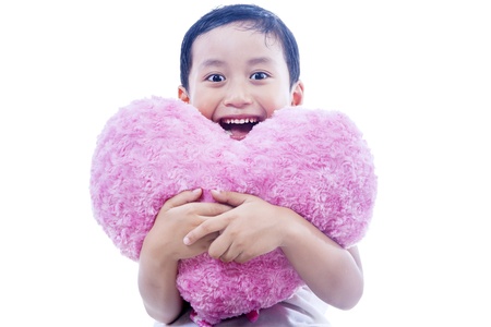 Asian boy is smiling cheerfully while holding a pink heart shape pillow on white backgroundの写真素材
