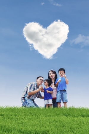 Family is having fun in the park while blowing bubbles with children under heart shape cloudsの写真素材