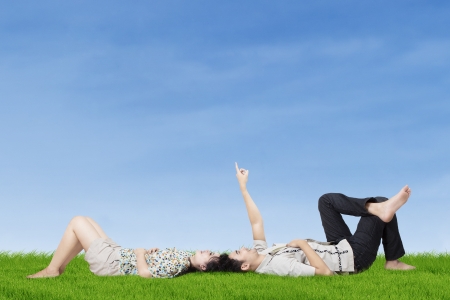 Happy young couple lying on the grass while pointing to copy space in the skyの写真素材