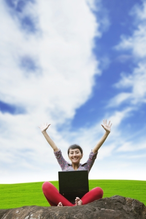 Asian woman raising her hands while using laptop in the park under blue  skyの写真素材