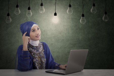 Beautiful female muslim has idea under light bulbs while working with her computerの写真素材