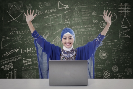 Happy female muslim raised her arms with laptop on written boardの写真素材