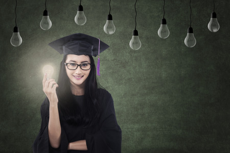 Beautiful Asian female graduate holding lit bulb under lampsの写真素材