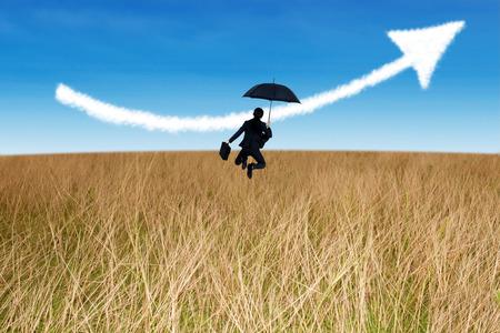 Young businesswoman jumping holding umbrella in field and arrow up signの写真素材