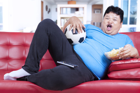 Overweight man sitting lazy on sofa while drinking beer and eat snackの写真素材