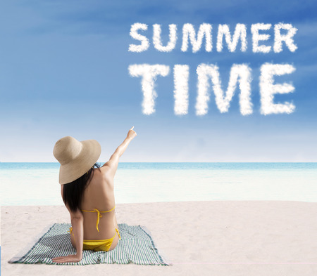 Portrait of smiling beautiful woman wearing bikini sitting on mat and pointing at a text of summer timeの写真素材