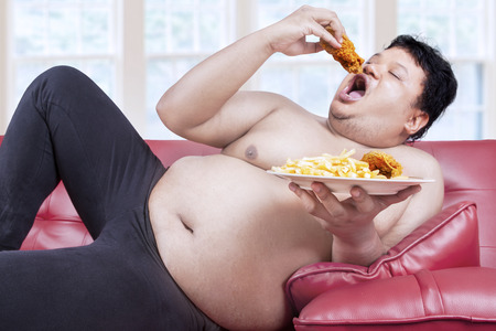 Fat man eats fast food while sitting on sofa at home