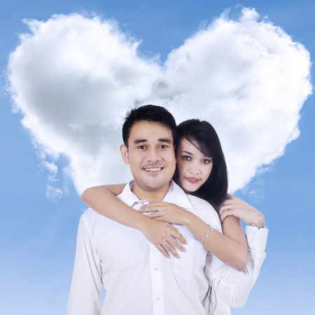 Happy young couple standing under clouds shaped of heartの写真素材