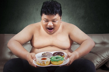 Cheerful fat man looking at donuts while sitting on the sofa at homeの写真素材