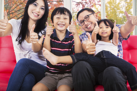 Joyful family sitting on sofa and giving thumbs up at camera with autumn background on the windowの写真素材