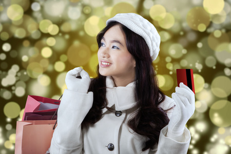 Portrait of young girl in winter coat holding credit card and shopping bags with a light glitter backgroundの写真素材