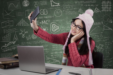 Lovely teenage girl sitting in the classroom while wearing warm clothes and taking picture with a...