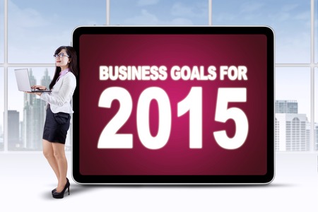 Young businesswoman using laptop computer while daydreaming and leaning on a board of business goals...