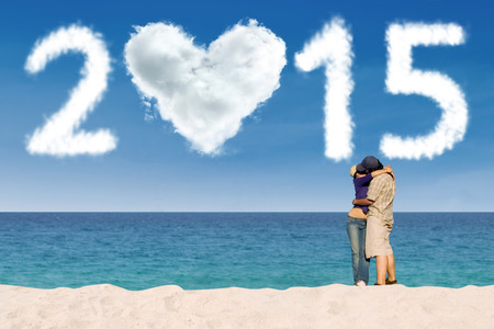 Young couple standing on beach, embracing and kissing under cloud shaped number 2015