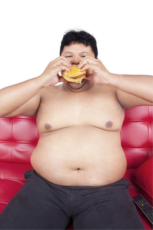 Portrait of asian man with fat body sitting on sofa while eating burgerの写真素材