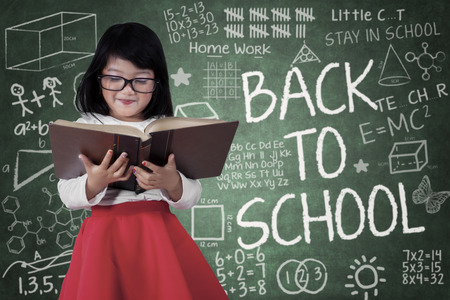 Portrait of little student standing in front of blackboard while reading a book with a text of back to schoolの写真素材