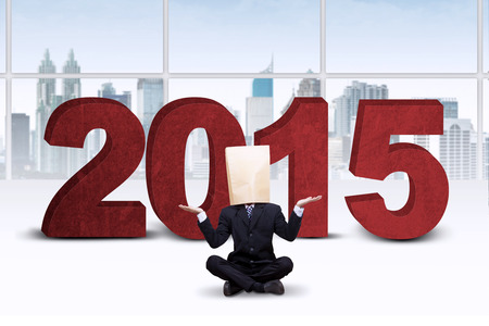 Confused businessman with a paper bag on head, sitting in front of numbers 2015 in office