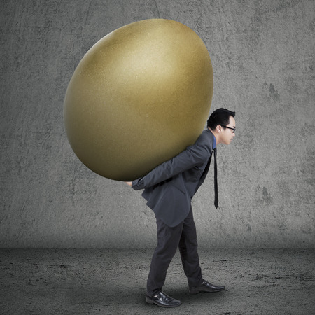 Young asian businessman carrying a golden egg as his profit, shot on gray backgroundの写真素材