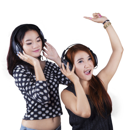 Portrait of two beautiful teenage girls dancing while listening music with headphones, isolated over whiteの写真素材