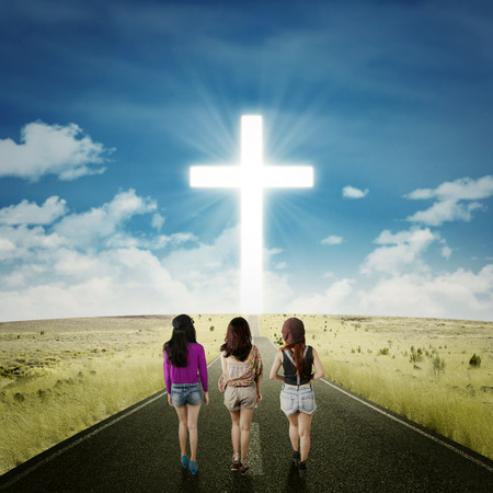 Rear view of three teenager girls walking together on the road toward a cross on the end of the roadの写真素材