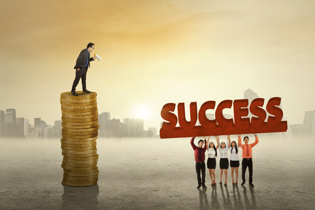 Young business leader standing on the stack of golden coins while giving order to his employeesの写真素材