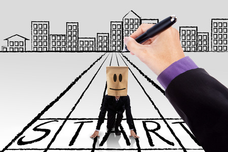 Male entrepreneur with a cardboard head kneeling on a start line with a hand giving a guide to startの写真素材