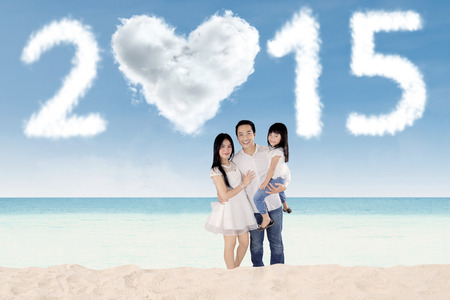 Portrait of happy family smiling at camera while standing on beach under cloud shaped number 2015