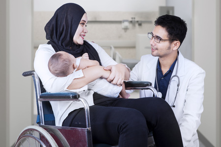 Male doctor talking with young mother and her baby sitting on the wheelchair at hospitalの写真素材