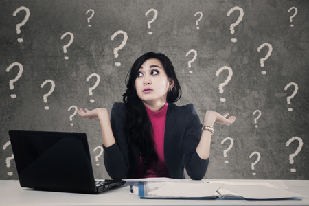 Beautiful businesswoman sitting in the office and looks confused with question marks on the backgroundの写真素材
