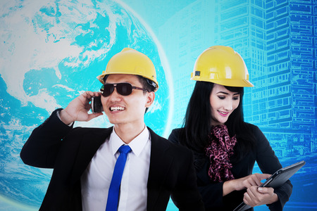 Two young engineer using cellphone and tablet to communicate, shot with world map in futuristic interfaceの写真素材