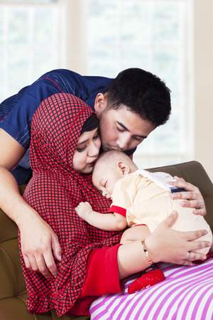 Portrait of two young muslim parents kiss their baby boy on the sofa at homeの写真素材