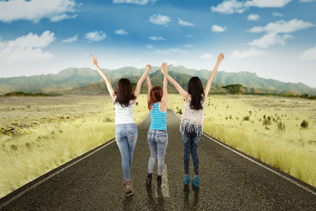Rear view of three teenage girls walking on the countryside road and raised hands togetherの写真素材
