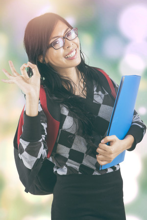 Young modern student smiling on the camera while showing okay sign, shot with bokeh backgroundの写真素材