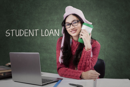 Female student sitting in classroom while holding a hot drink and wearing knitted clothes, laptop on the table and a text on the blackboardの写真素材