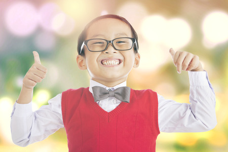 Portrait of little schoolboy showing thumbs up on the camera, shot with light glitter backgroundの写真素材