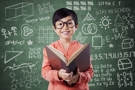 Portrait of pretty little girl standing near the chalkboard in the class while reading a bookの写真素材