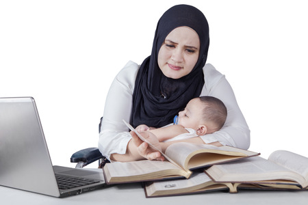 Portrait of young woman working with textbooks on desk while carrying newborn babyの写真素材