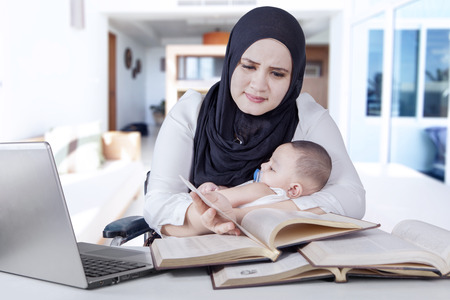 Portrait of disabled woman sitting on wheelchair while holding her baby and working on deskの写真素材