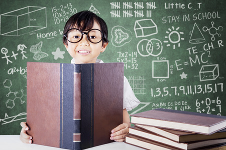 Lovely female primary school student wearing glasses and reading textbooks in the classroomの写真素材