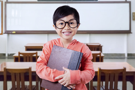 Cheerful female elementary school student smiling at the camera while holding a book in the...