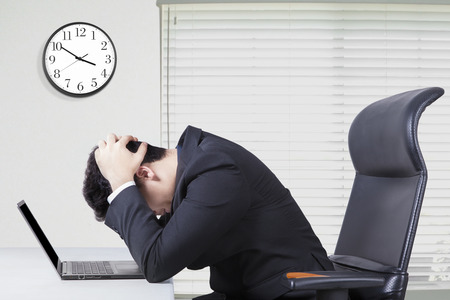 Young entrepreneur sitting in the office and looks confused with a laptop on desk and clock on the wallの写真素材